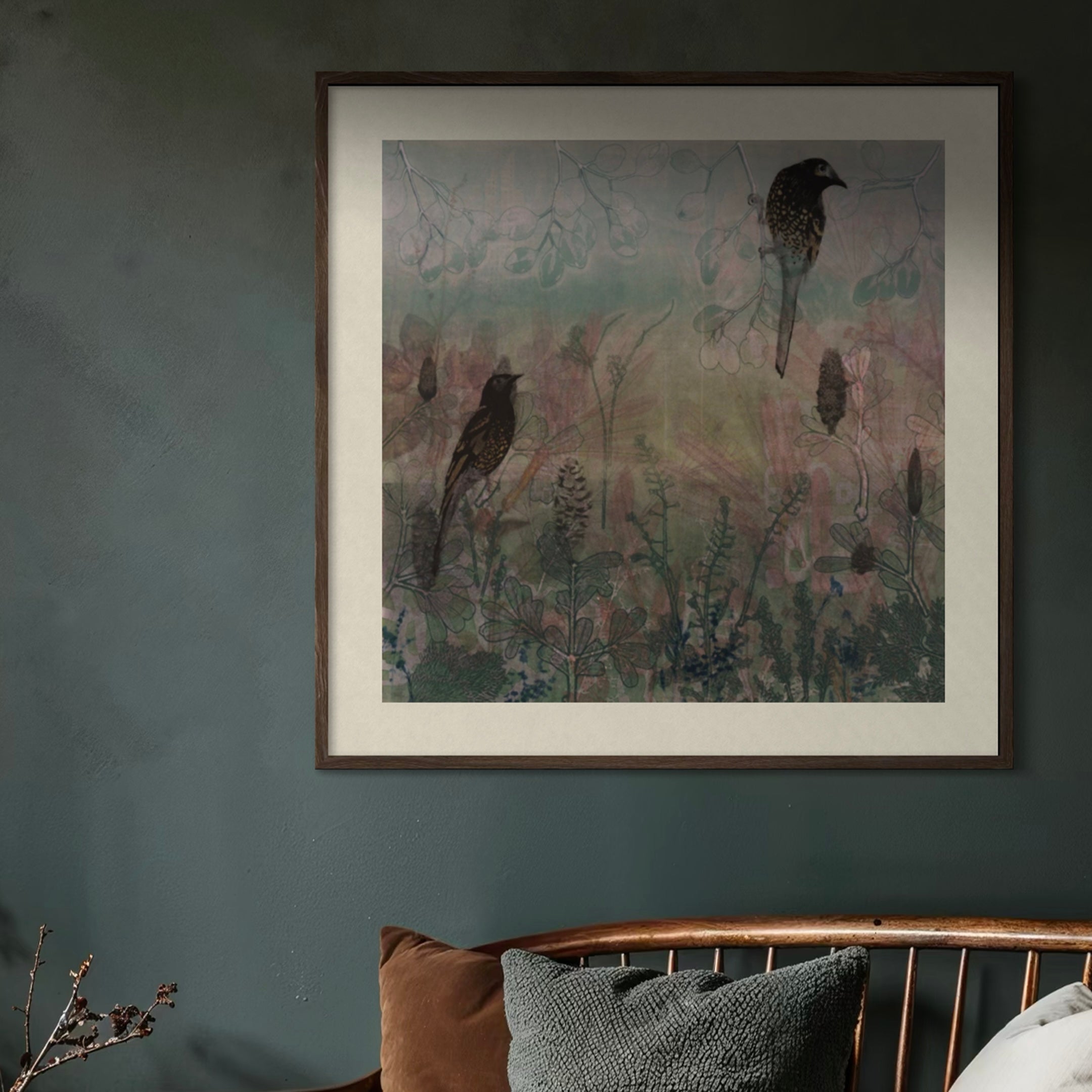 A framed wall art print featuring two dark birds over a soft, muted landscape in dusty blue, pink, and green tones, displayed on a dark textured wall above a wooden chair with cushions.